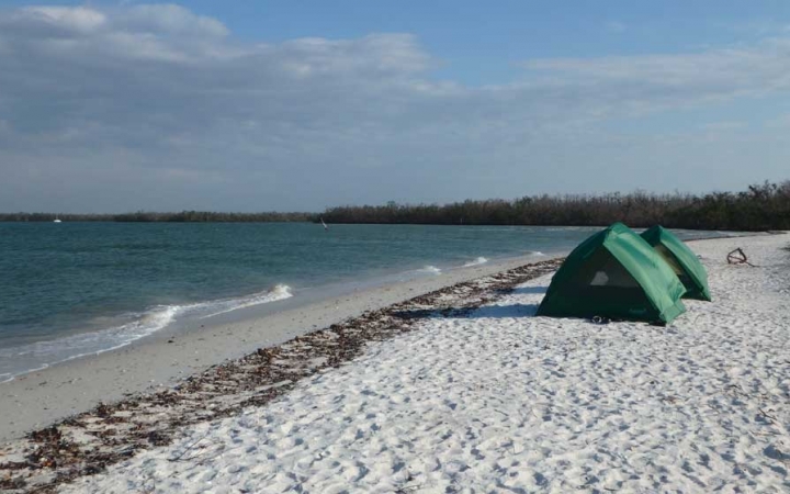 Two tents rest on a sandy beach beside a blue body of water. 